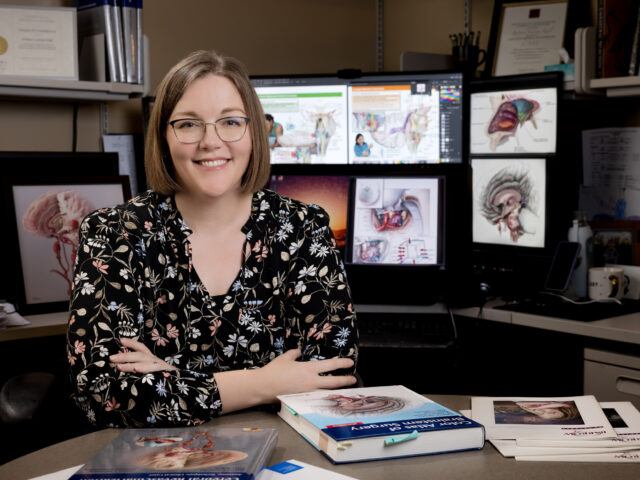 Kristen Larson Keil leans on her desk. Her artwork is displayed on computer screens behind her and on textbooks and journals in front of her.