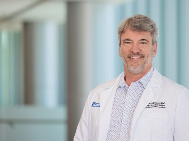 Dr. Robert Bowser is pictured in a brightly lit lobby of the Barrow Neuroplex. He is wearing a white lab coat and smiling at the camera.