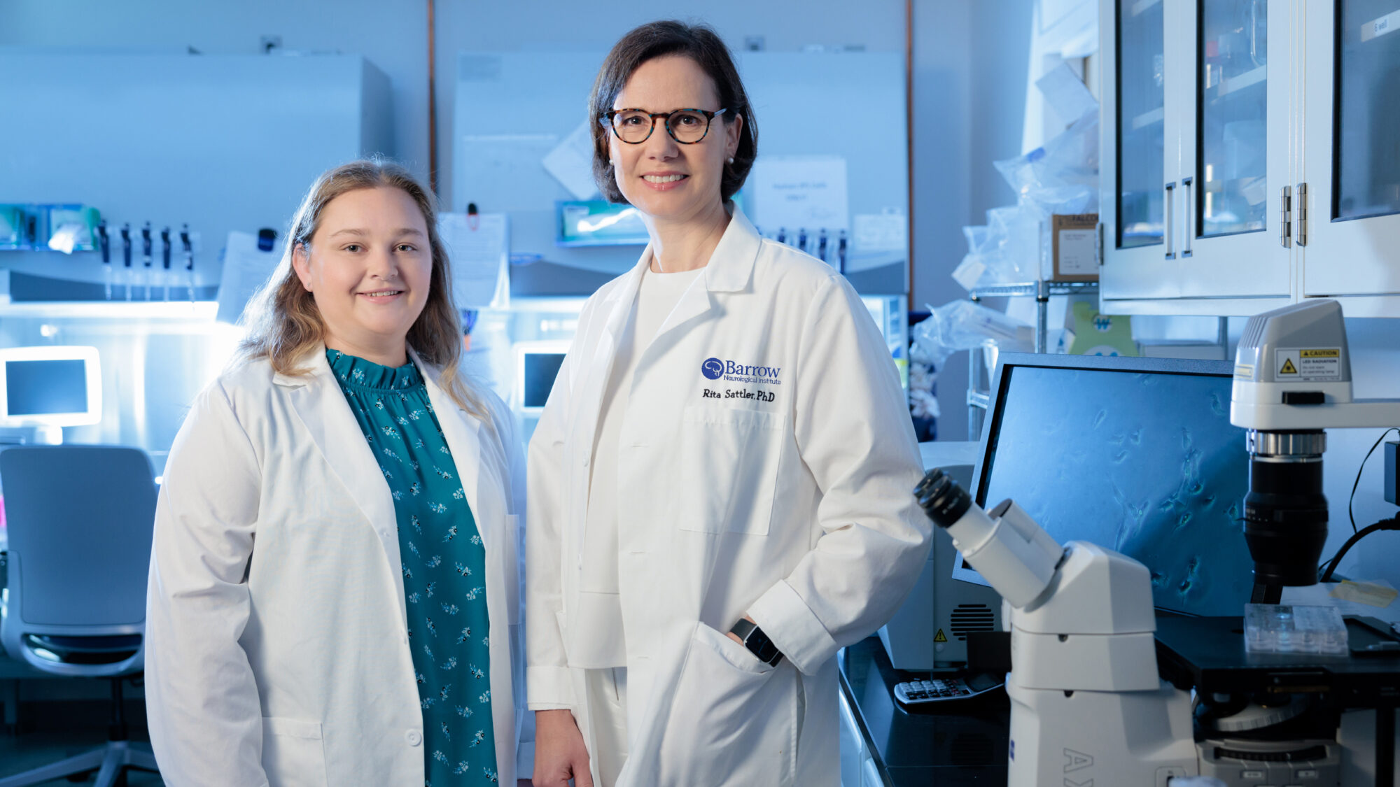 Briana Ondatje and Rita Sattler, PhD, pose for a photo in the Sattler Laboratory. They are both wearing white lab coats and smiling at the camera.
