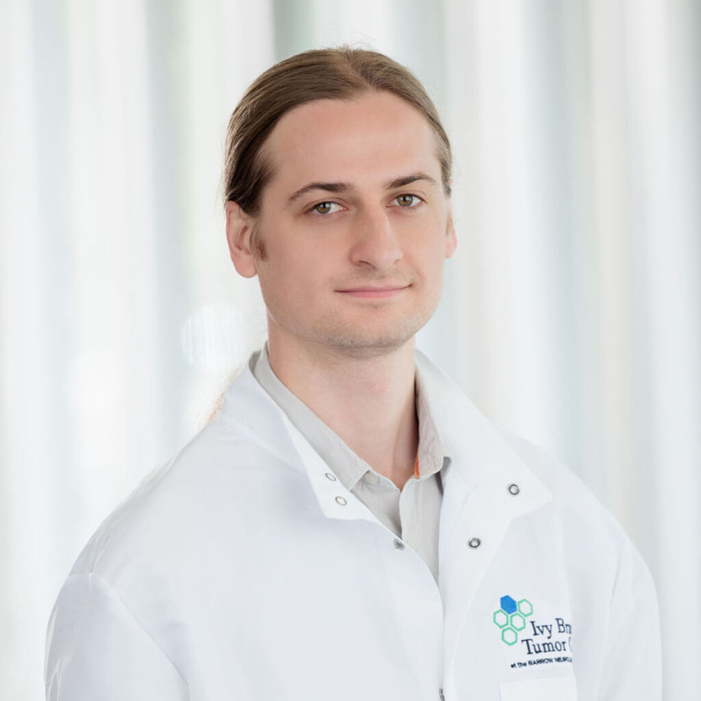 Portrait of William Knight. He is standing in a brightly lit hallway in the hospital and wearing a white lab coat with the Ivy Brain Tumor Center logo on the chest.