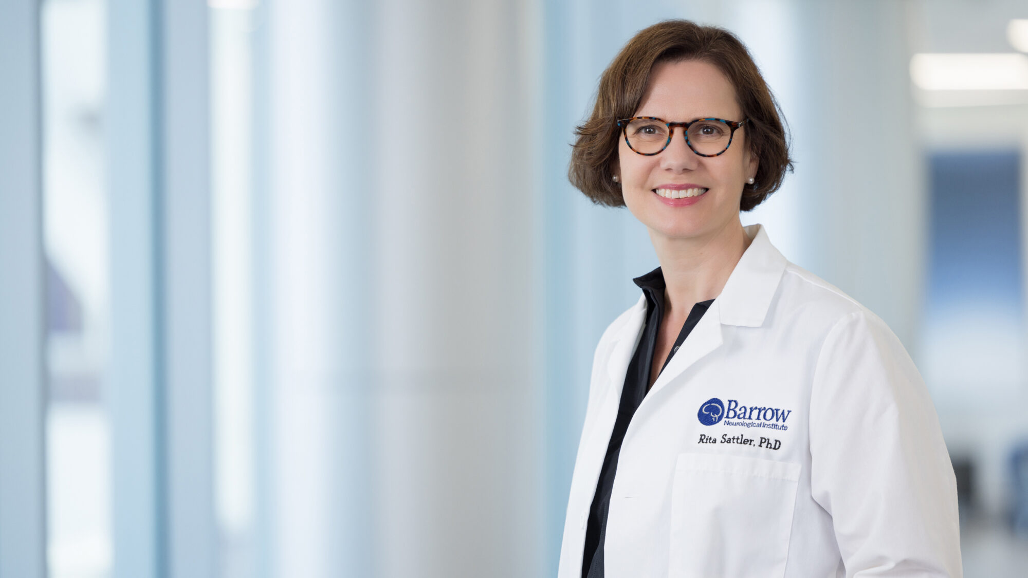 Portrait of Rita Sattler, PhD, in a brightly lit hallway at Barrow. She is wearing a Barrow-branded white lab coat and smiling at the camera.
