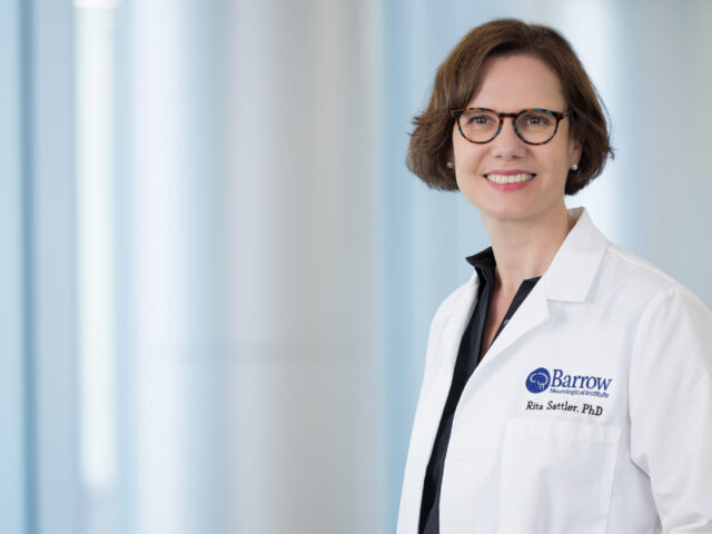 Portrait of Rita Sattler, PhD, in a brightly lit hallway at Barrow. She is wearing a Barrow-branded white lab coat and smiling at the camera.