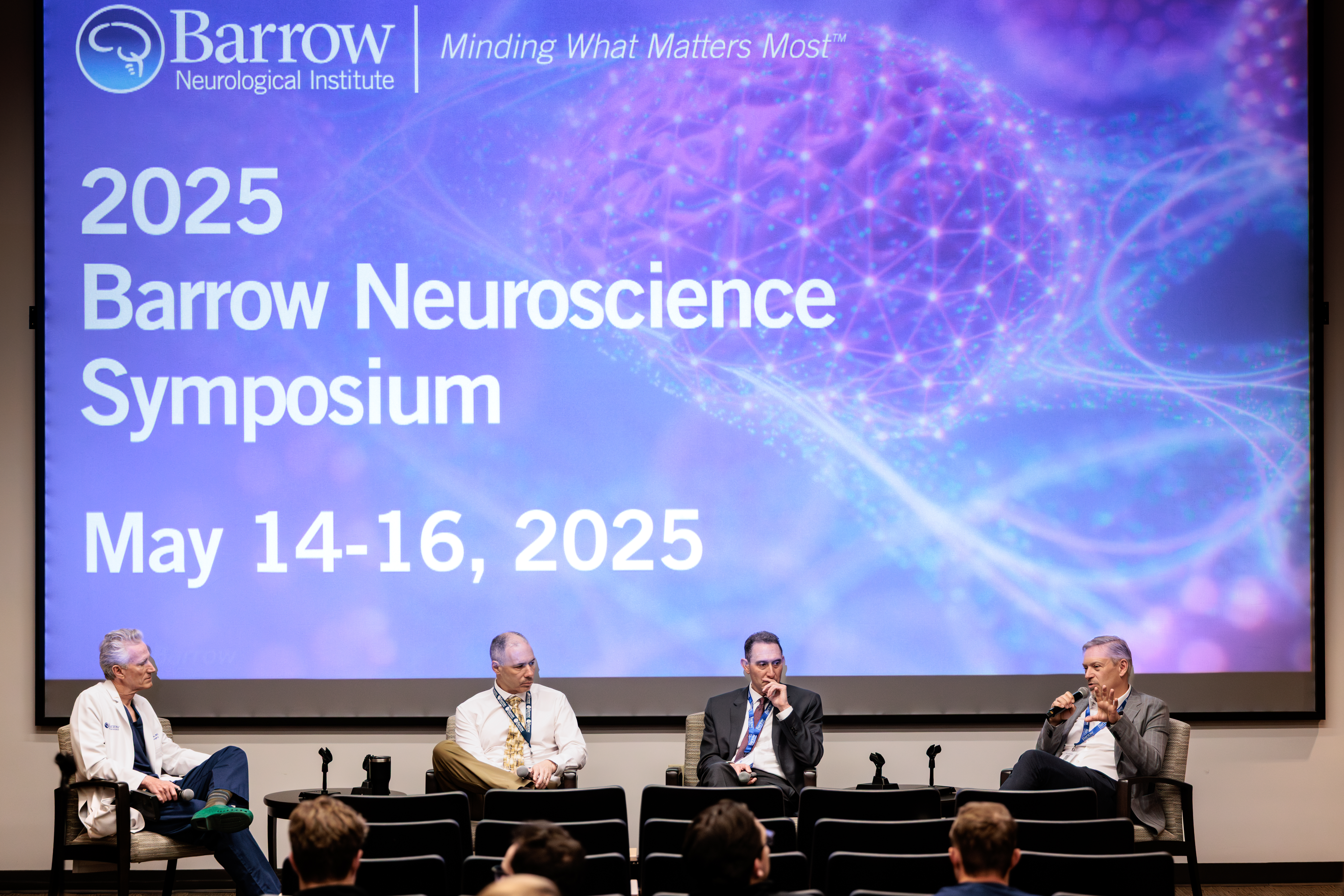 A panel of speakers seated in front of a screen in an auditorium. The screen says "2025 Barrow Neuroscience Symposium May 14-16, 2025" and shows an abstract illustration of a brain.
