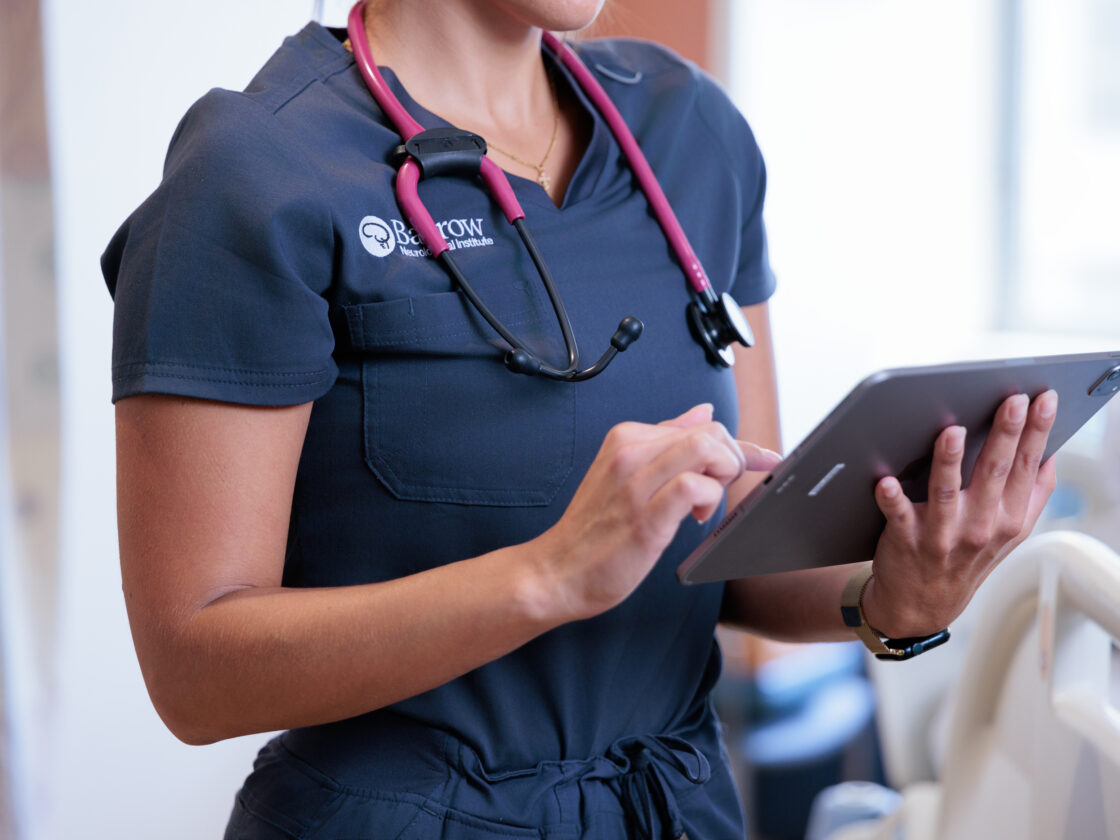 A barrow nurse in blue scrubs with a pink stethoscope enters information on a tablet in a hospital room.