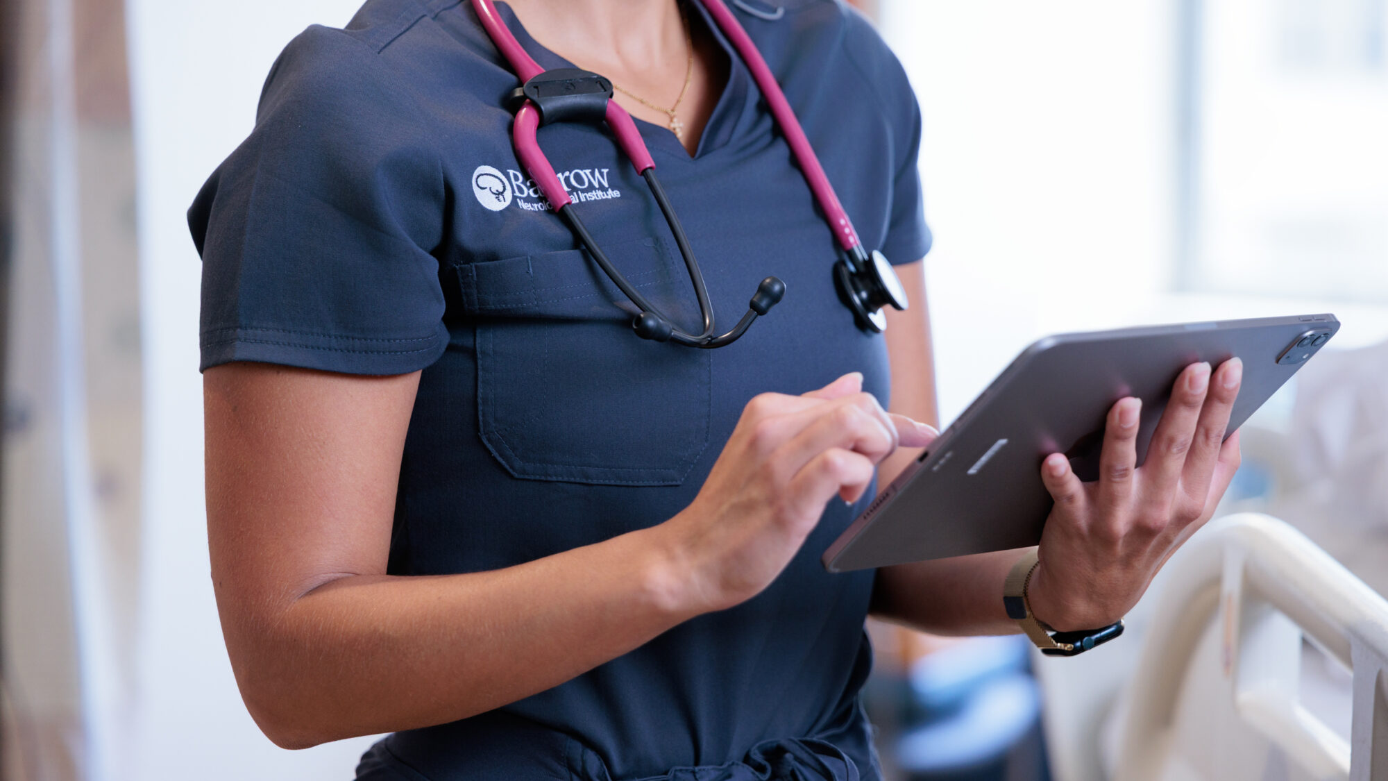 A barrow nurse in blue scrubs with a pink stethoscope enters information on a tablet in a hospital room.