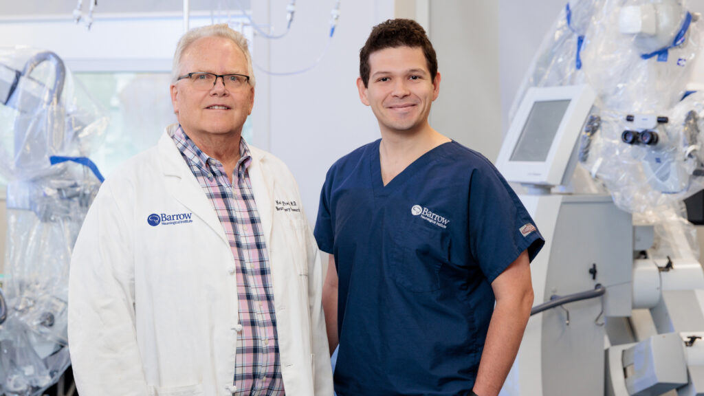 Dr. Preul and Dr. Yangi pose for a photo in Dr. Preul's lab. Dr. Preul is wearing a white lab coat, and Dr. Yangi is wearing navy blue scrubs. They are smiling at the camera.