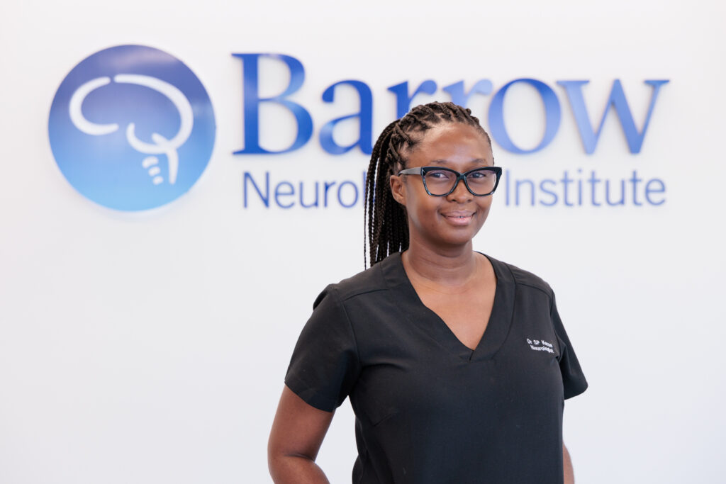 Sandane Chauke-Korae, MD, in front of a Barrow logo in a lobby of the hospital. She is wearing scrubs and smiling softly at the camera.