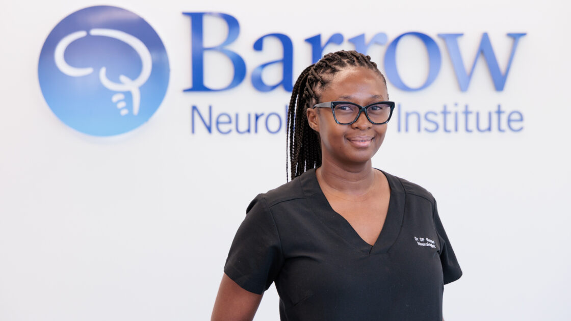 Sandane Chauke-Korae, MD, in front of a Barrow logo in a lobby of the hospital. She is wearing scrubs and smiling softly at the camera.