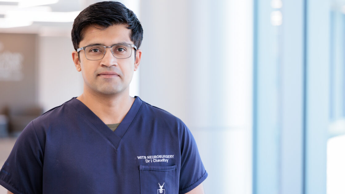 Irtiza Chaudhry, MD, in a brightly lit lobby at Barrow. He is wearing scrubs and smiling softly at the camera.