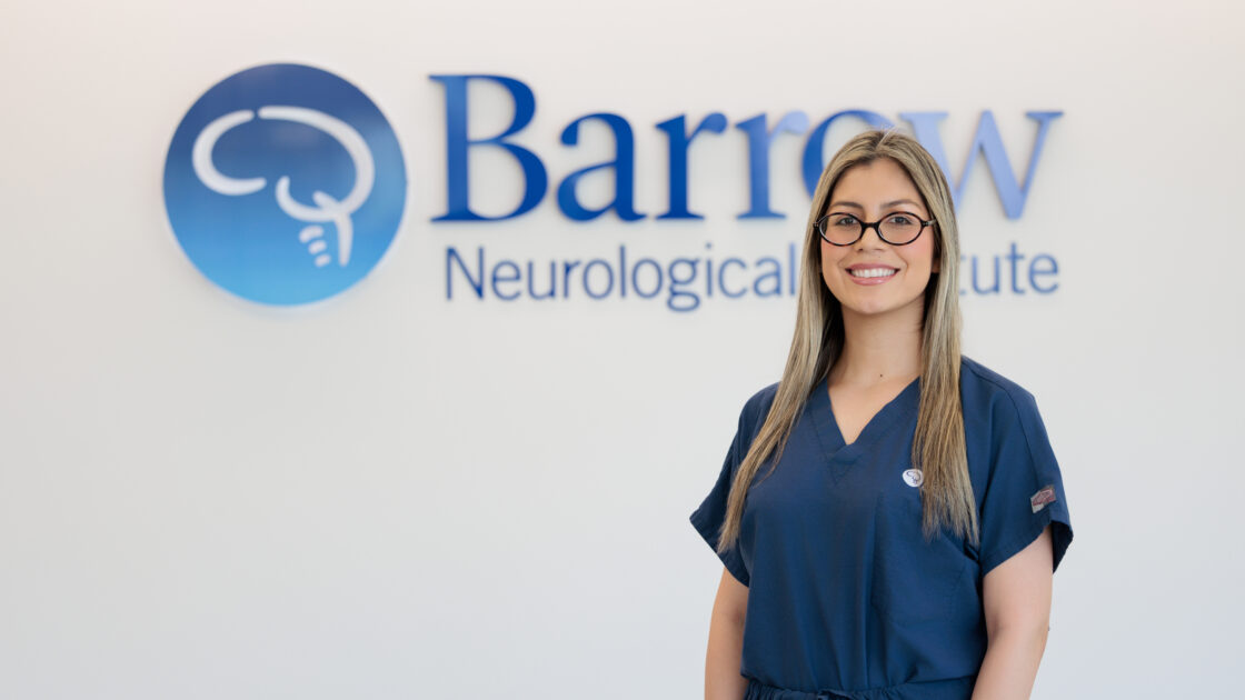 Portrait of Dr. Beatriz Galeano in front of the Barrow logo in a brightly lit lobby. She is wearing Barrow-branded scrubs and smiling at the camera.