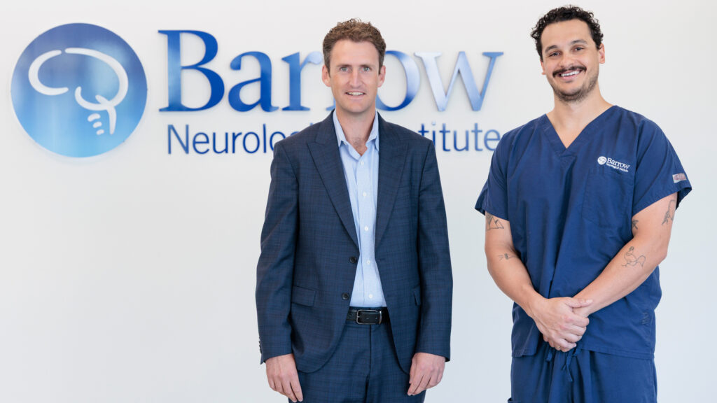 Dr. Rory Murphy and Dr. Benjamin Rodriguez stand in front of the Barrow logo in a brightly lit lobby of the hospital. Dr. Murphy is wearing a suit, and Dr. Rodriguez is wearing Barrow-branded scrubs.