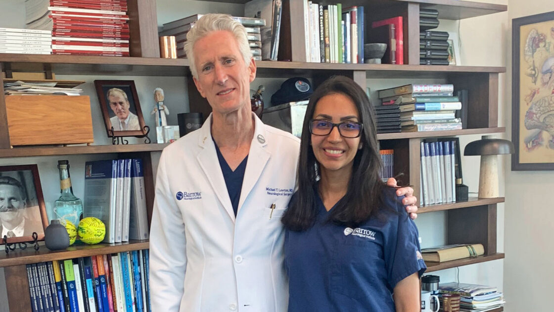 Photo of Drs. Viana and Lawton in front of a bookshelf in Dr. Lawton's office. The two are smiling at the camera. Dr. Lawton is wearing a white lab coat, and Dr. Viana is wearing blue Barrow-branded scrubs.