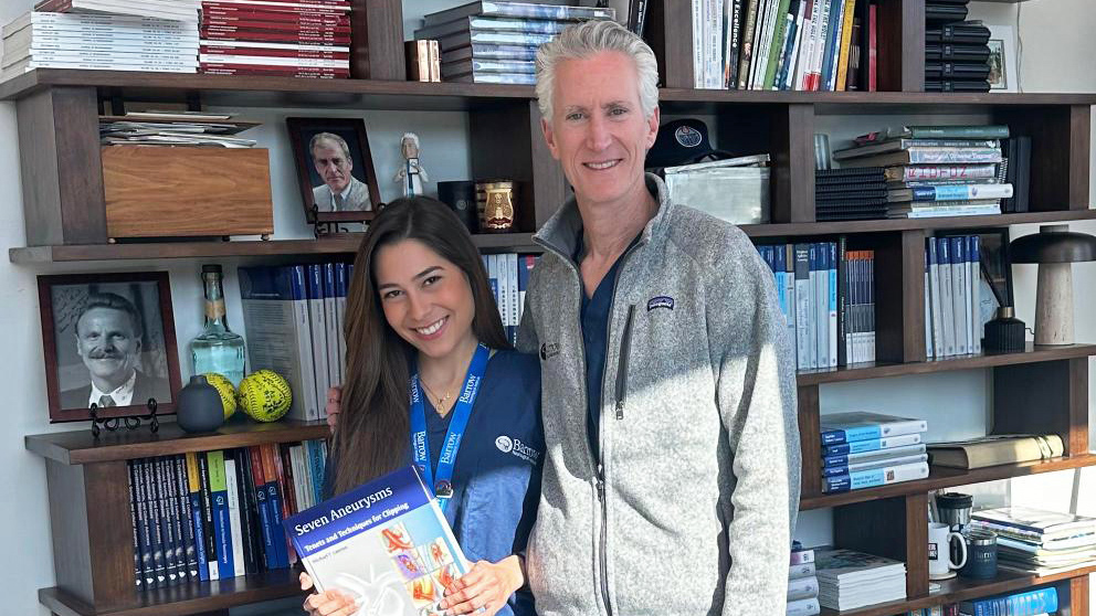 Dr. Marina Mendes Melo and Dr. Lawton pose in front of the bookshelf in his office. Dr. Mendes Melo is holding up Dr. Lawton's "Seven Aneurysms" textbook.