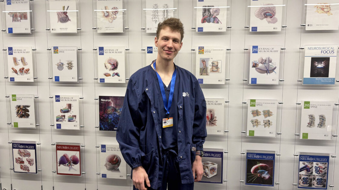 Photo of Karl Kapahnke in front of a wall of journal covers illustrated by the Barrow Neuroscience Publications Department. Karl is wearing blue Barrow scrubs.