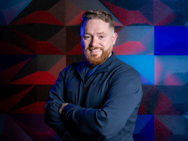 Luke O'Neill poses in front of multicolored panels. His arms are crossed, and he is smiling.