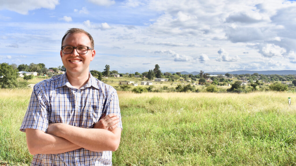 Barrow neurology resident Ryan Wagner, MD, poses for an environmental portrait in South Africa. He is in front of a field, sparsely populated with structures. He has his arms folded and is smiling at the camera.