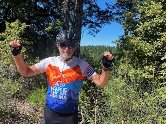 Dr. Jeremy Shefner holds up his fists in celebration of his bike ride. He is wearing a jersey that says "Bike the US for MS."