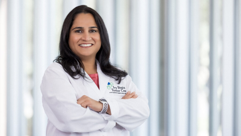 Shwetal Mehta, PhD, poses for a photo in a lobby at Barrow Neurological Institute. She is smiling at the camera with her arms folded and wearing a white lab coat with the Ivy Brain Tumor Center logo.