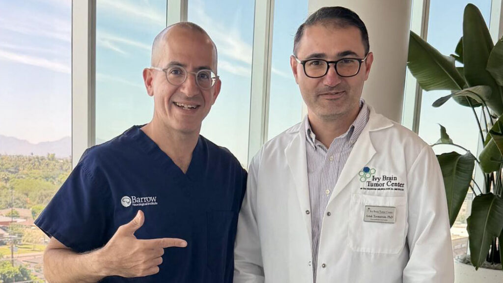 Artak Tovmasyan, PhD, and Nader Sanai, MD, in a brightly lit lobby at the Ivy Brain Tumor Center. Dr. Sanai is pointing to Dr. Tovmasyan.