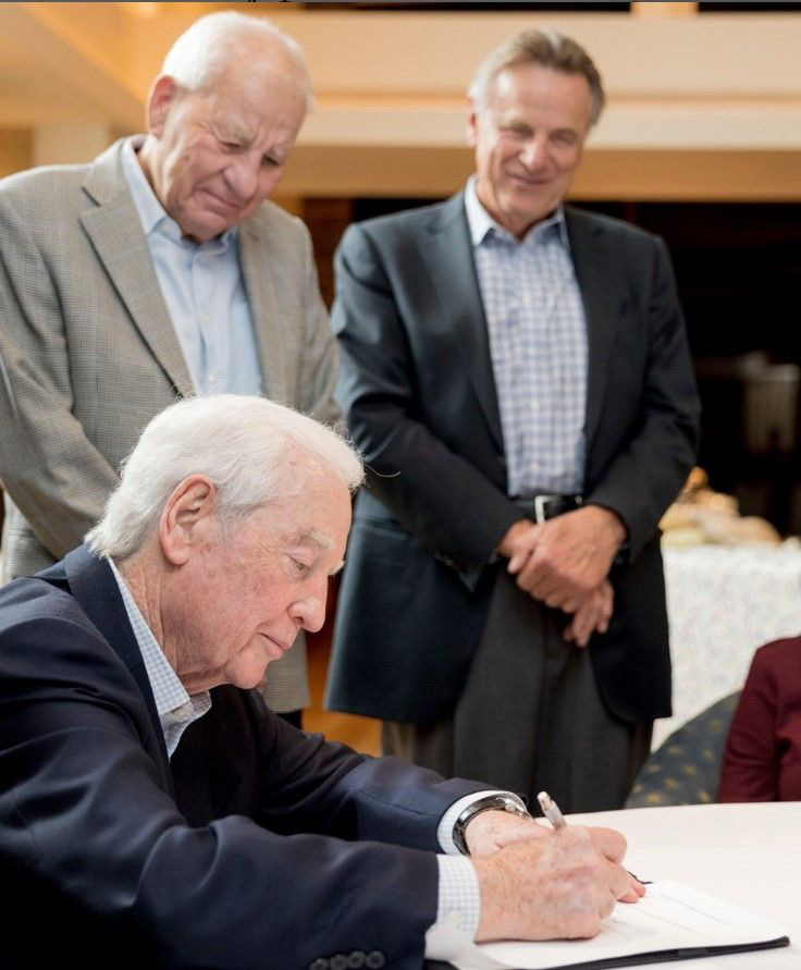 A photo of Bill Franke signing a document to create the Franke Global Neuroscience Education Center. Dr. Robert Spetzler and Dr. Volker Sonntag are standing next to him.