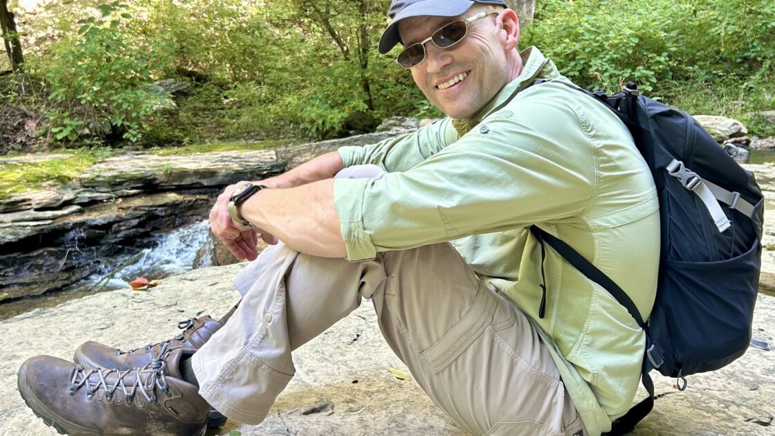 Ivy Brain Tumor Center patient Jeff Muse sitting on a rock wearing a backpack in hiking attire in front of a forest full of green trees.