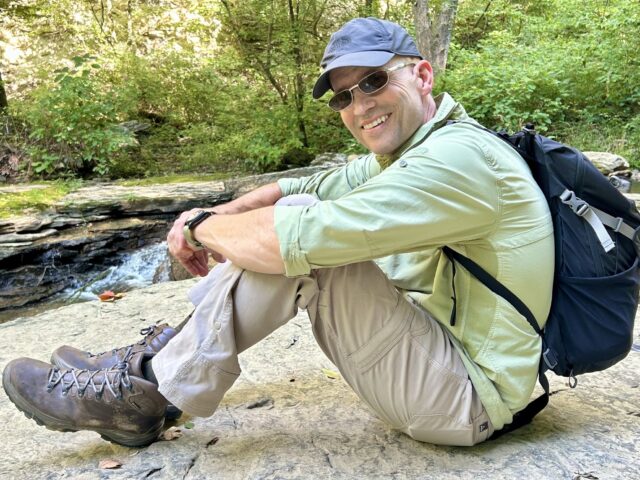 Ivy Brain Tumor Center patient Jeff Muse sitting on a rock wearing a backpack in hiking attire in front of a forest full of green trees.